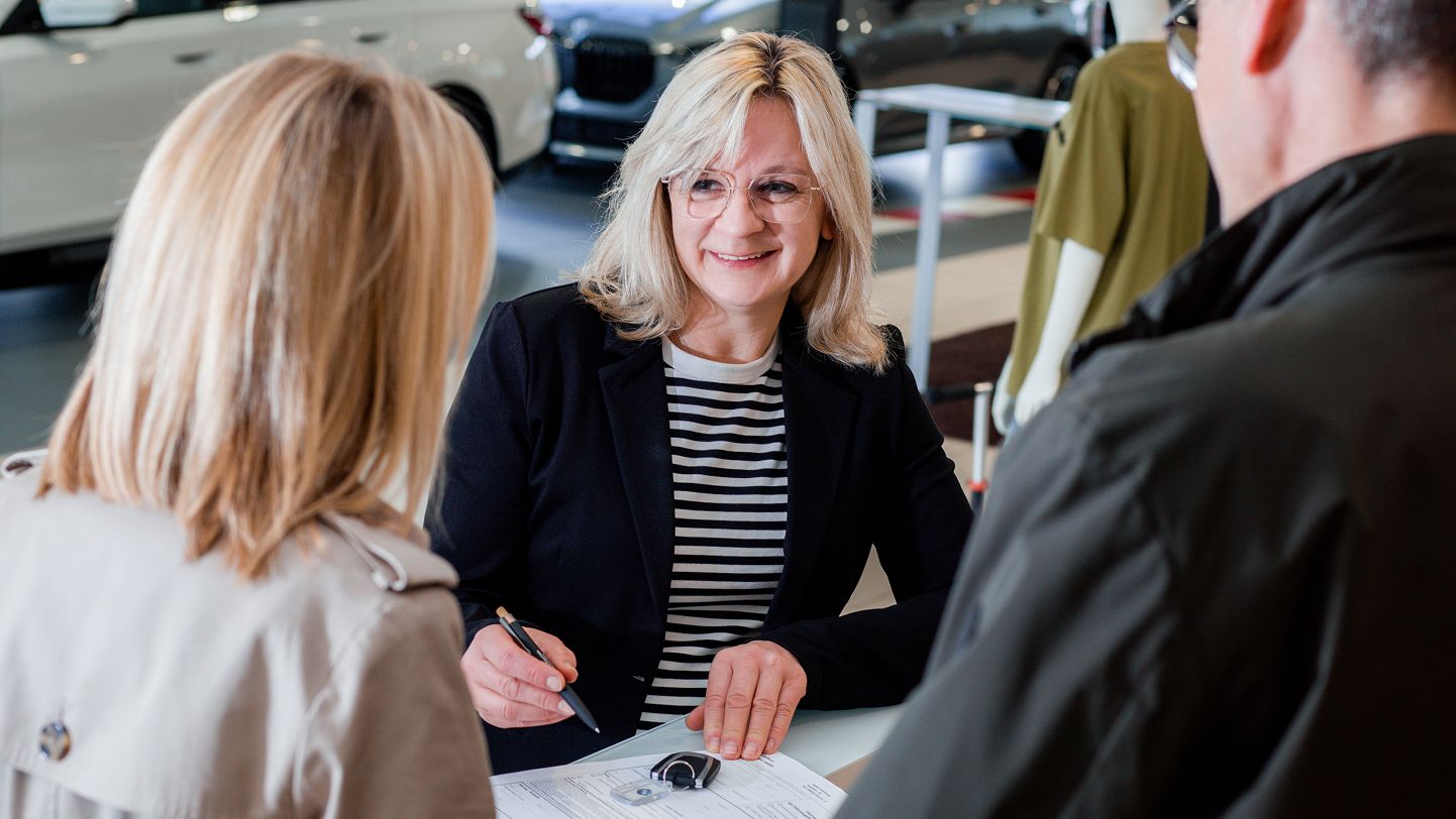 Serviceberaterin im Autohaus berät ein Paar am Schreibtisch, zeigt Vertragsunterlagen und legt Autoschlüssel bereit, moderne Fahrzeuge im Hintergrund.