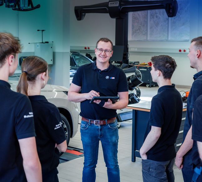 BMW-Techniker mit Tablet unterweist vier Auszubildende in moderner Kfz-Werkstatt, Diagnosegerät und Fahrzeuge im Hintergrund.
